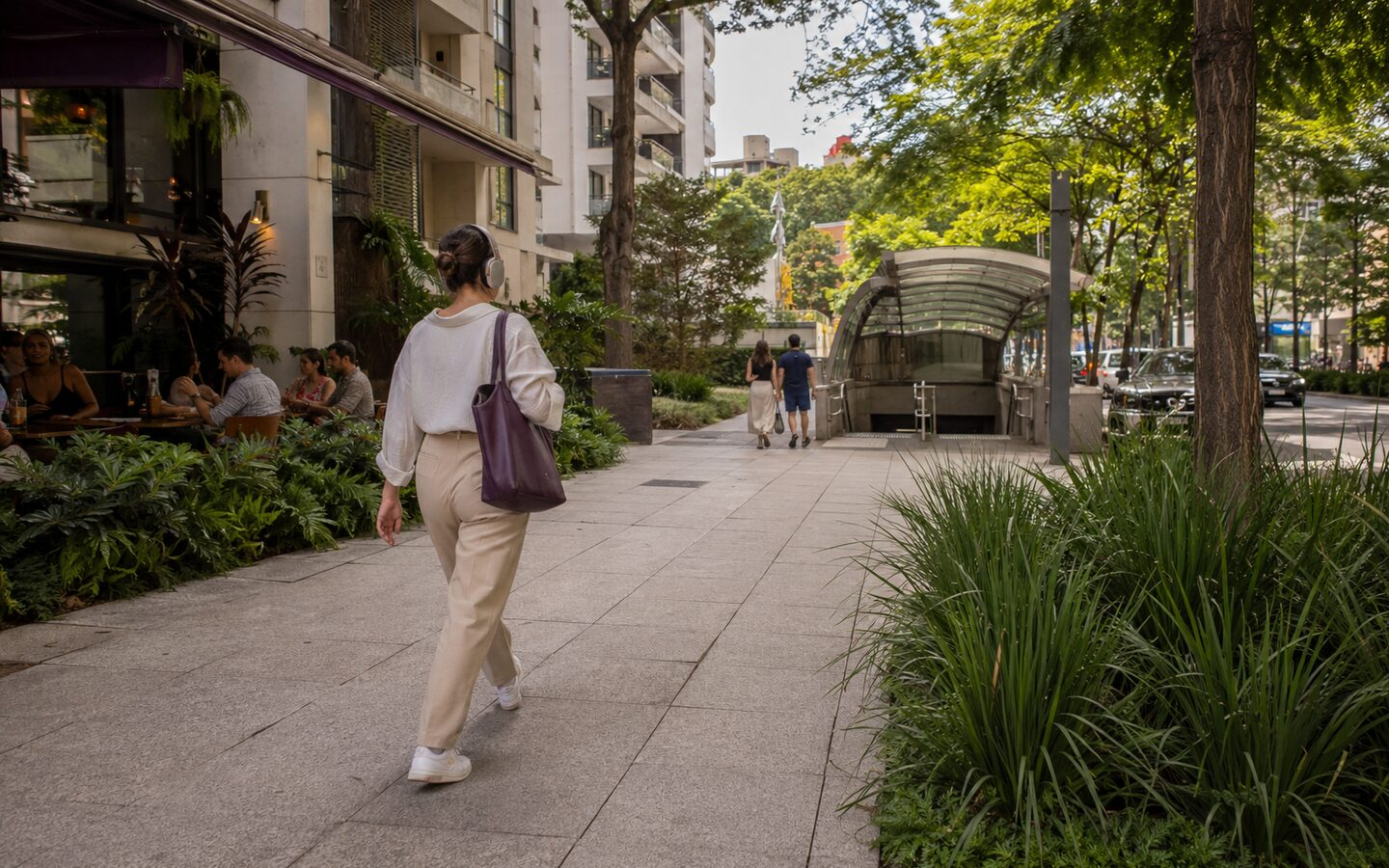 Moradora caminhando por calçada arborizada de Moema em direção a uma estação de metrô